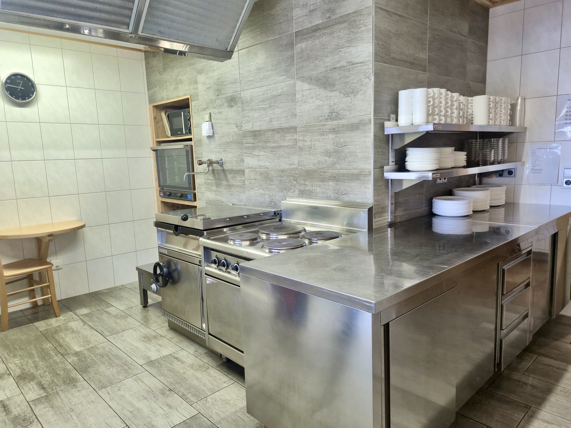 Moderne Edelstahl-Gastroküche im Selbstversorgerhaus Maishofen mit großem Kochfeld, Arbeitsinsel und Regalen mit Geschirr.